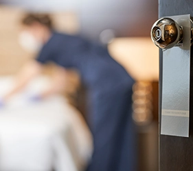 Hotelreinigung - Im Vordergrund ist ein goldener Türknauf zu sehen und im Hintergrund eine Frau in einem Hotelzimmer, die ein Bett bezieht - Clean Team Solingen